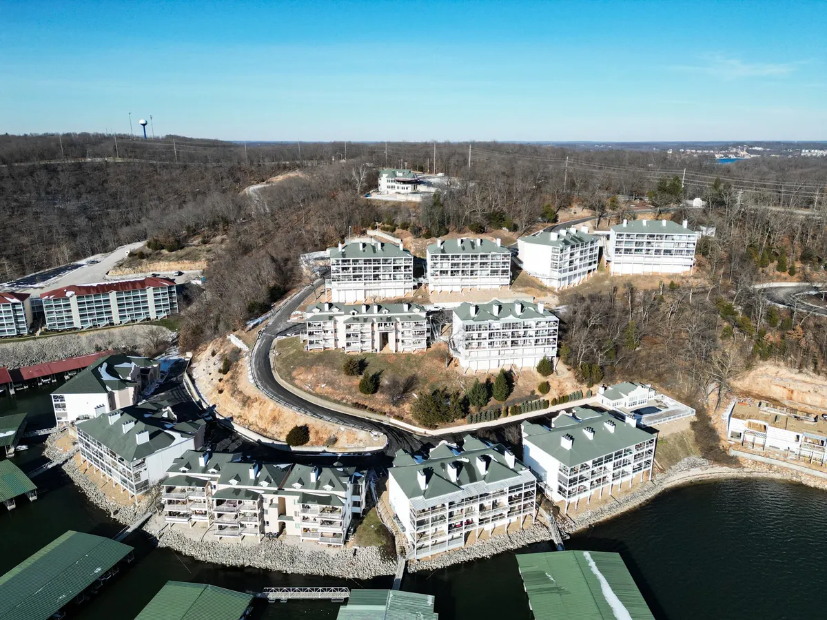 This image captures waterfront buildings nestled along the lakeshore, surrounded by lush trees and a winding road. The tranquil water and shoreline are clearly visible, evoking the essence of lakeside living with easy access to boating and fishing.