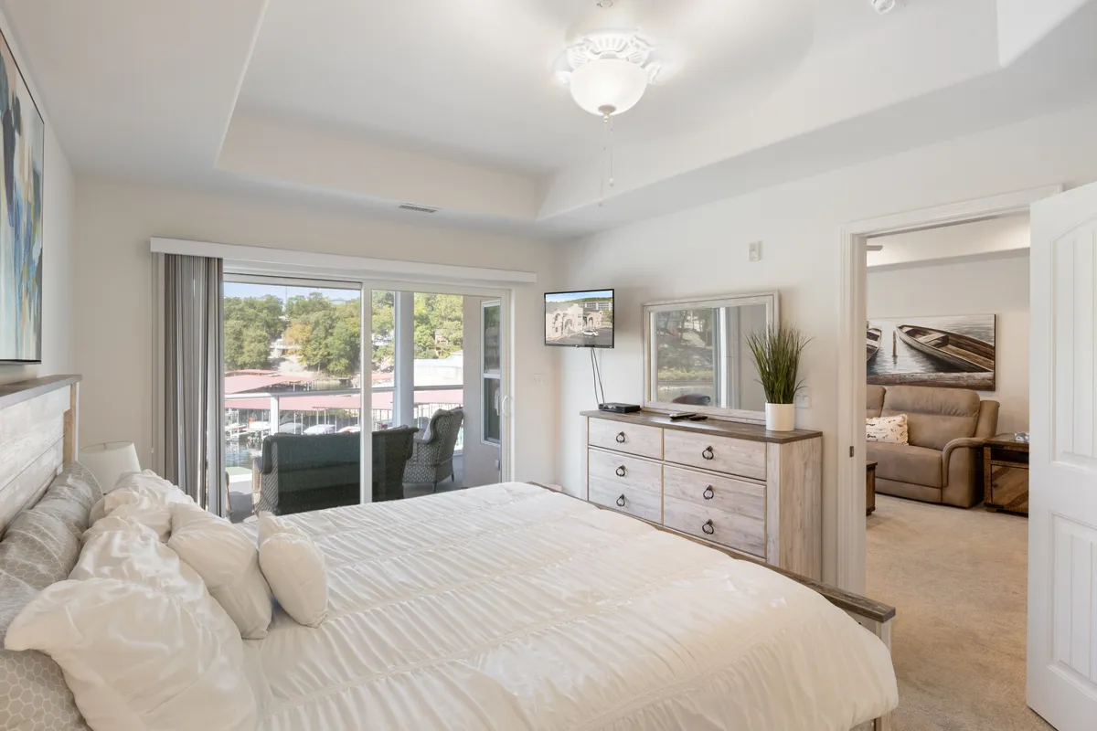 This image showcases a serene bedroom featuring a bed with crisp white linens, a large window with a sliding door that likely offers access to the deck, a dresser with a mirror, and a television on the wall. Soft natural light fills the room, creating an inviting atmosphere for relaxation and restful nights.