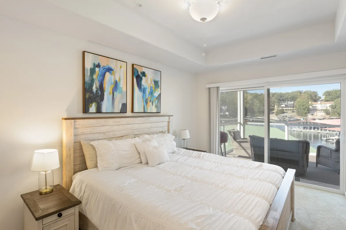 A bright, peaceful bedroom with white bedding, tasteful artwork on the wall, and a sliding glass door leading to a balcony. The spacious room is flooded with daylight, emphasizing a tranquil retreat ideal for winding down.