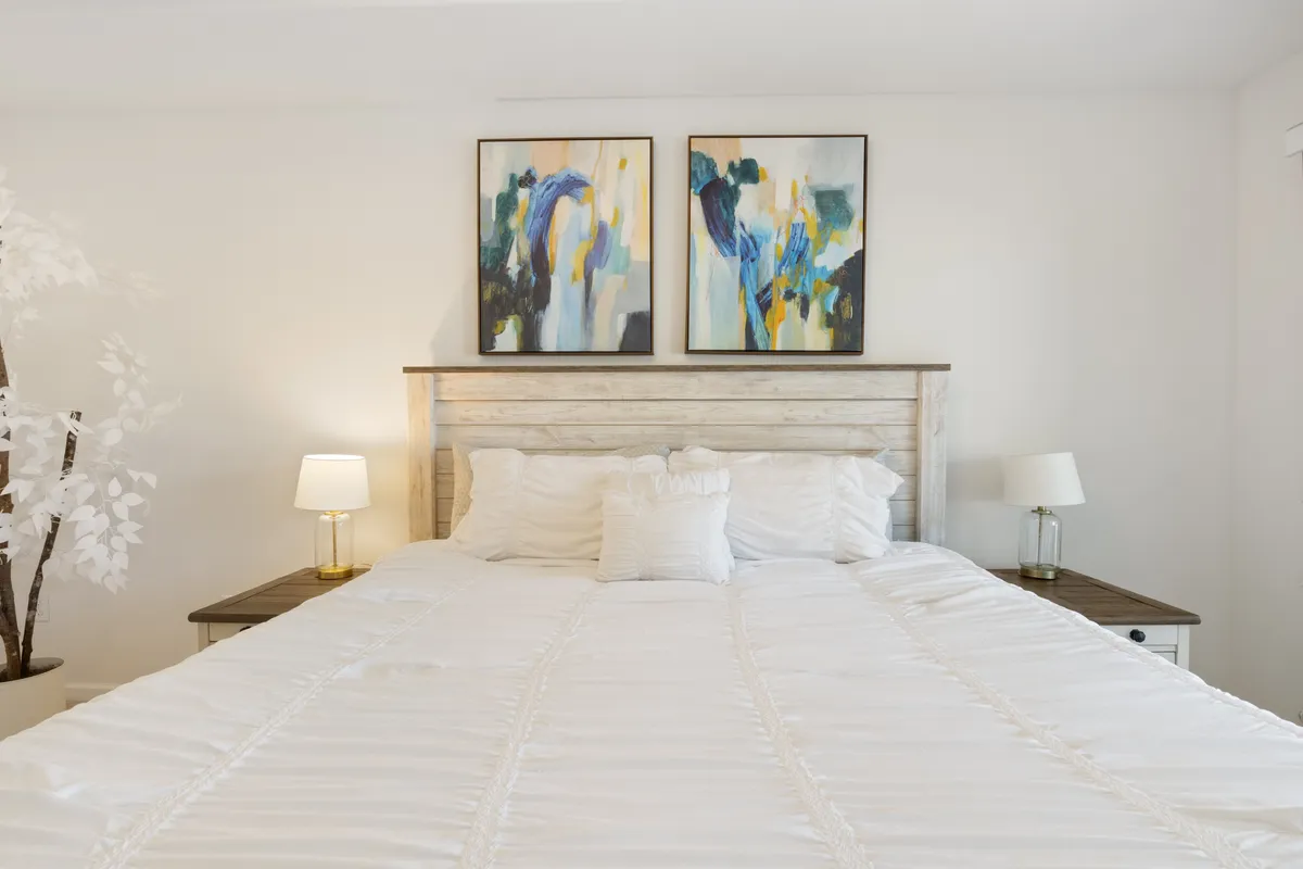 A tranquil bedroom with white bedding, decorative pillows, and gentle accent lighting from bedside lamps. Artwork and a decorative plant create an inviting, cozy environment ideal for restful nights.