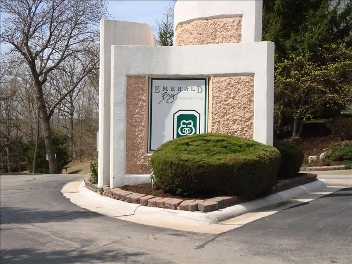 A photo of the Emerald Bay community sign, framed by landscaped bushes and mature trees. The scene signals arrival at a friendly, well-maintained lakeside neighborhood—suggesting both a sense of place and security.