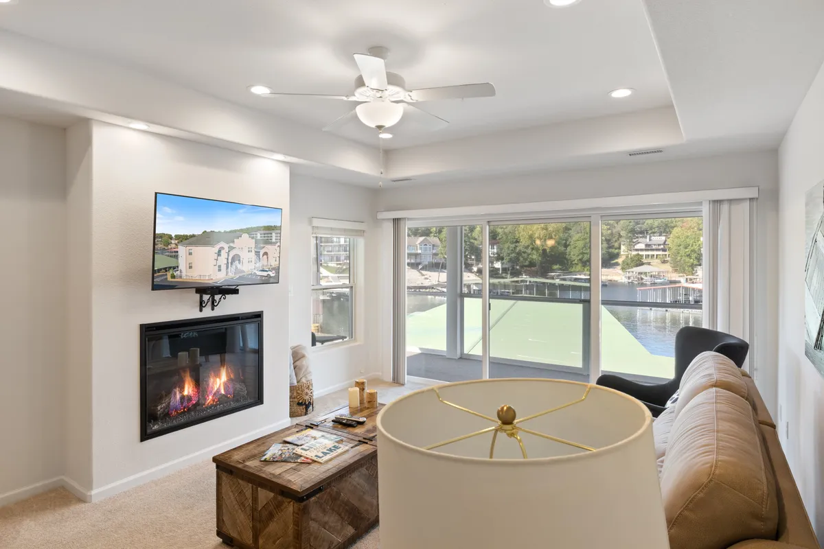 A spacious living room featuring plush sofas arranged around a fireplace, with a large TV and coffee table. Expansive windows frame stunning views of the lake, filling the room with natural light and offering a tranquil backdrop. The ambiance is inviting, ideal for relaxation and social gatherings.
