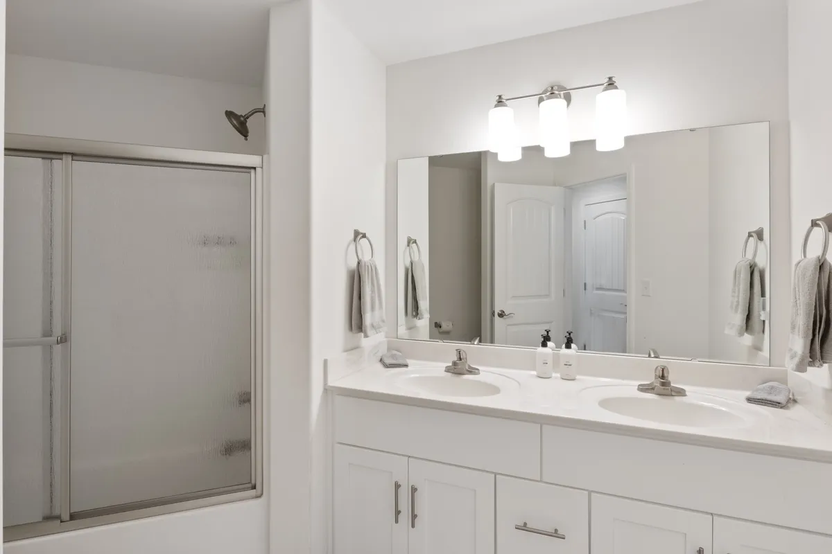 A modern bathroom featuring a double sink vanity, a large mirror, and a shower area. The setting is fresh and inviting, providing a glimpse of the convenience and comfort available for morning and evening routines.