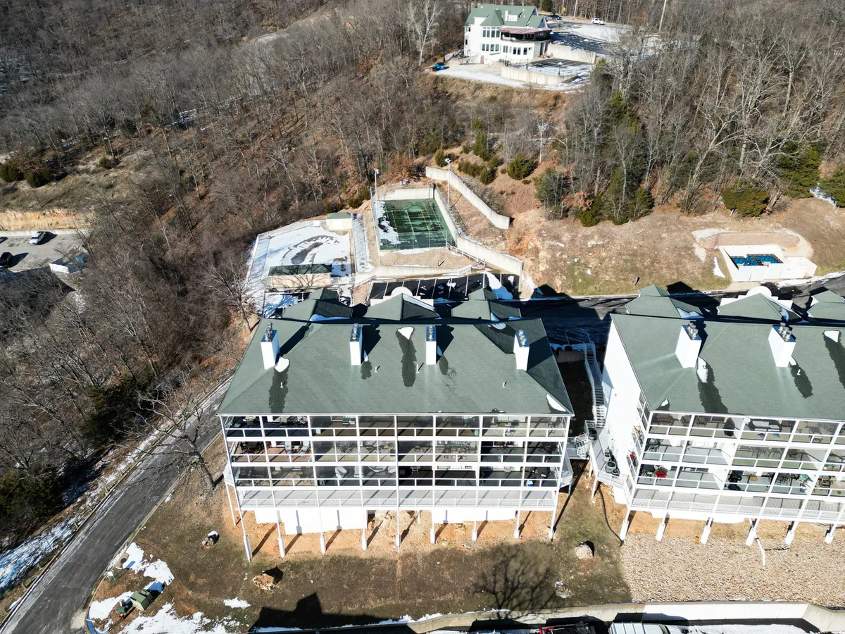 This image features the multi-story building complex with an inviting outdoor pool area surrounded by lush, wooded landscape. The pool, sun loungers, and gentle hill in the background evoke a resort-style ambiance and highlight the scenic outdoor amenities available to guests.