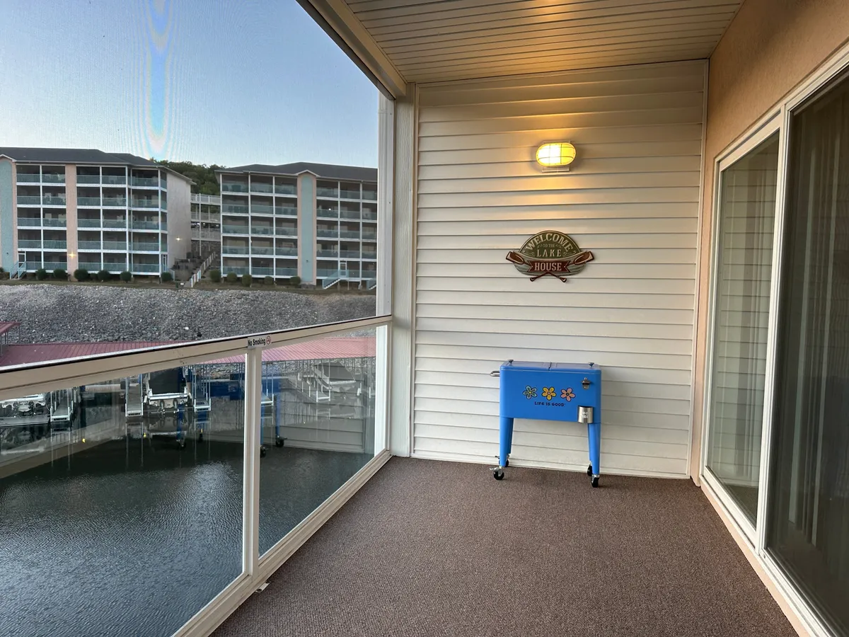 A scenic balcony view overlooking the lake, featuring a blue cooler, a welcome sign, and outdoor seating. The setting is perfect for relaxing, enjoying the sunset, or sharing a drink with friends and family, blending indoor comfort with outdoor beauty.