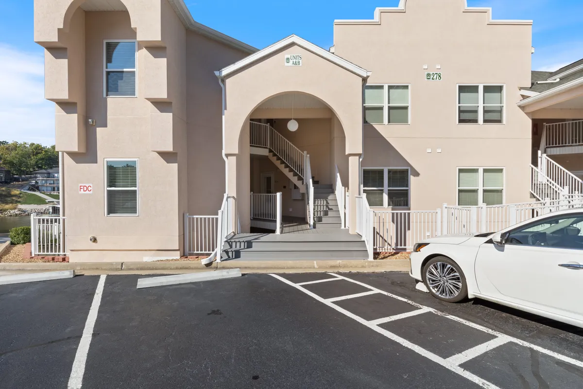 Shows the arched entrance to the condo building, a welcoming staircase, a parked white car, and landscaped surroundings. The scene conveys arrival, accessibility, and a sense of community.
